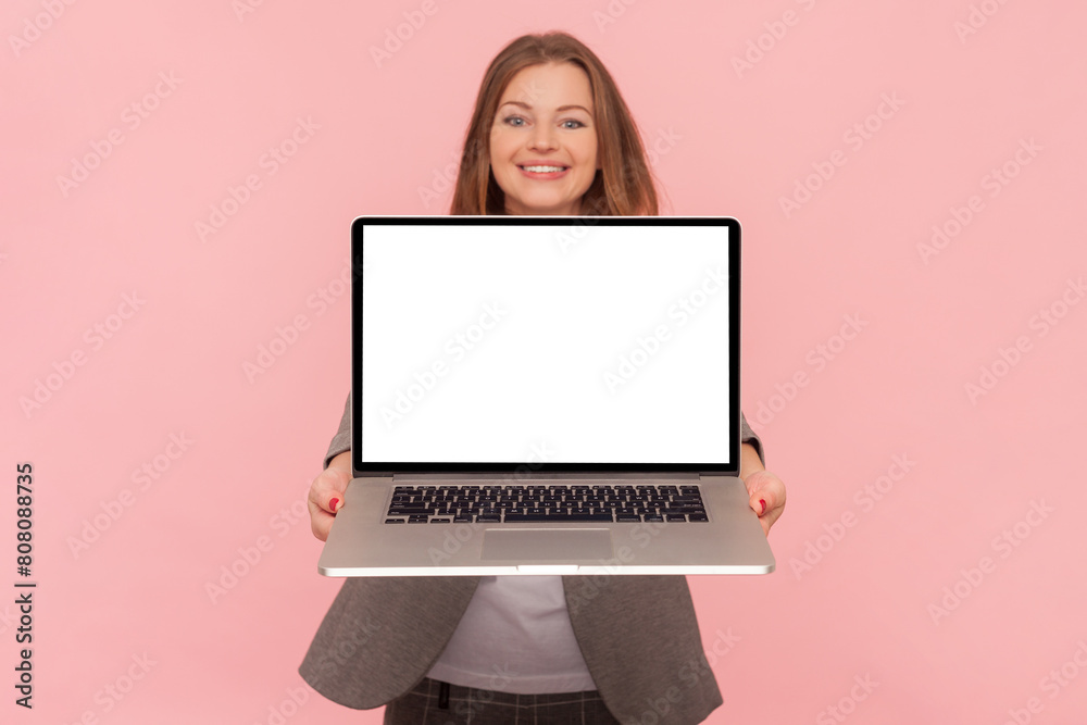 Portrait of smiling joyful woman with brown hair holding laptop with empty display, copy space for advertisement, wearing business suit. Indoor studio shot isolated on pink background. Portrait of smiling joyful woman with brown hair holding laptop with empty display, copy space for advertisement, wearing business suit. Indoor studio shot isolated on pink background.