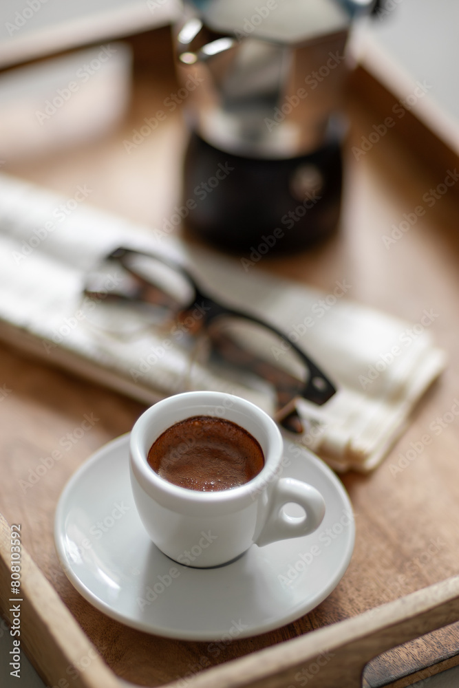 A freshly brewed espresso sits on a wooden tray with a pair of glasses and newspaper, inviting a relaxing break. A freshly brewed espresso sits on a wooden tray with a pair of glasses and newspaper, inviting a relaxing break.