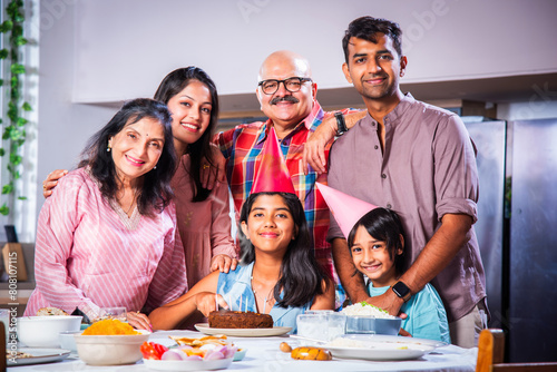 Indian asian grandparents with Family celebrating birthday by blowing candles on cake at home on dining table.