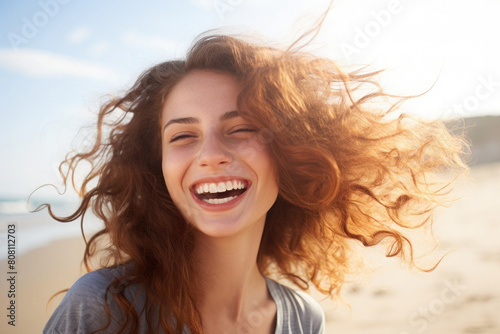 Beautiful young woman in gray shirt with curly red hair blowing in the wind, smiling brightly and laughing, enjoying on the beach as her hair is blowing in the wind.