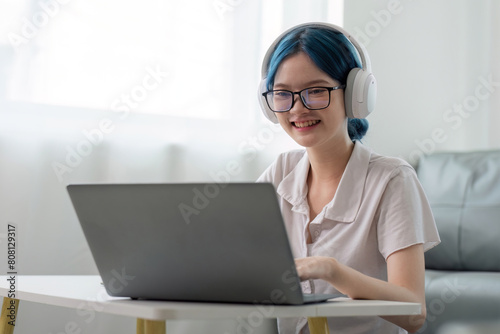 Smiling girl student wear headphone study online with teacher, happy young asian woman learn lecture watch webinar write notes look at laptop sit in home, distant education © Natee Meepian