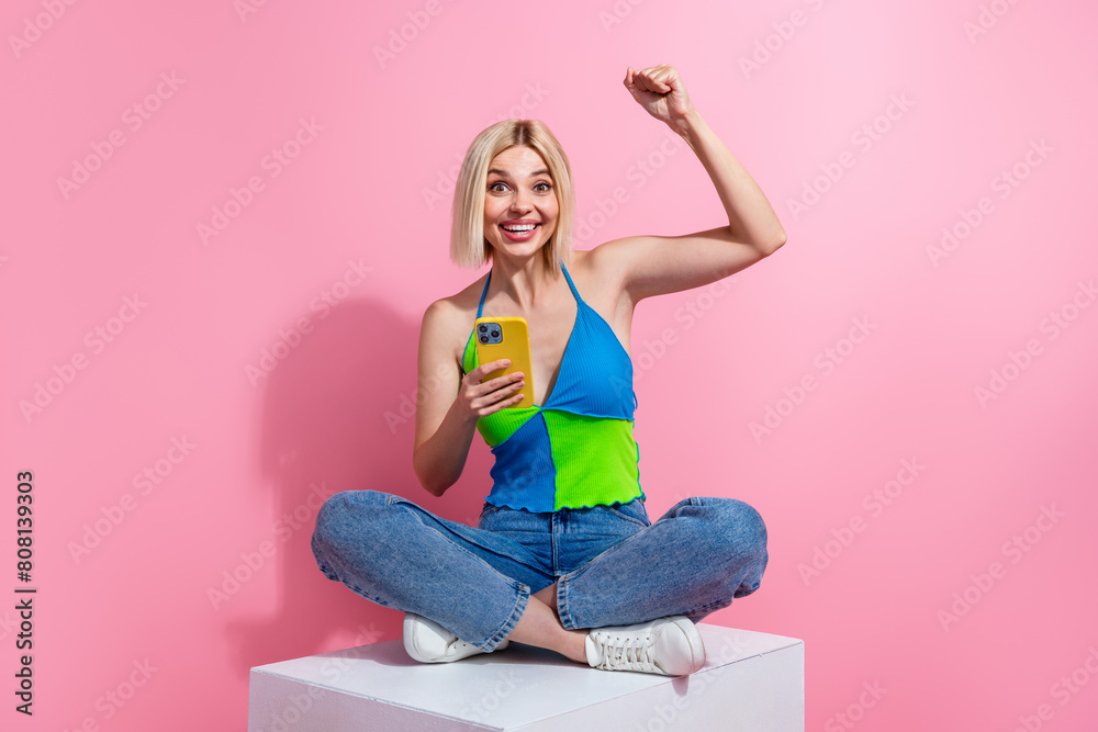 Full length photo of ecstatic girl dressed colorful top denim pants sit on cube hold smartphone win bet isolated on pink color background Full length photo of ecstatic girl dressed colorful top denim pants sit on cube hold smartphone win bet isolated on pink color background