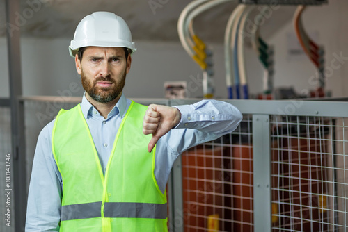 Sad man in a safety vest and helmet gives a thumbs down with negative expression, electrician engineer fail