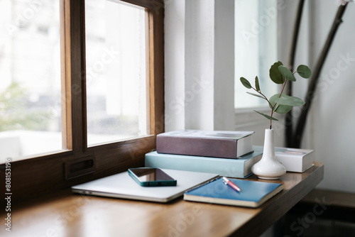 Comfortable workplace on wooden counter by the window and smart phone put on laptop, note book and pen, vase with small tree inside and a book in the background, Select focus on white vase