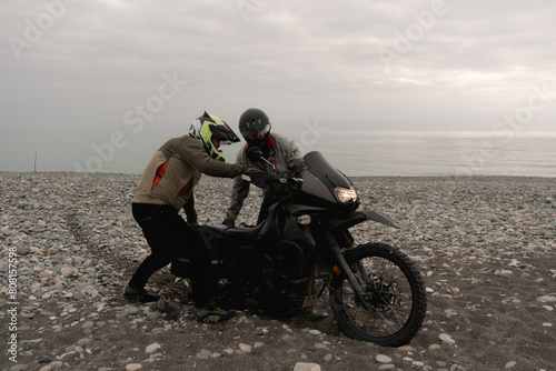 Friends bikers helping in trip on adventure motorcycle towing on sea shore pebbles