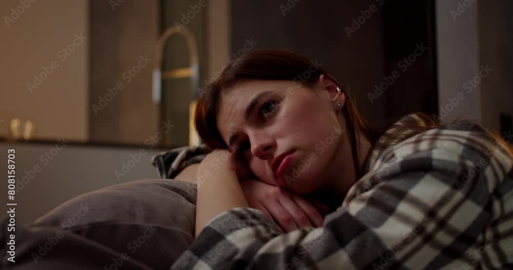 A calm and bored brunette girl in a checkered shirt leans on a pillow on a gray sofa she is bored and looks away in a modern apartment in the evening A calm and bored brunette girl in a checkered shirt leans on a pillow on a gray sofa she is bored and looks away in a modern apartment in the evening