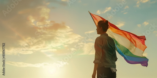 Pride Month concept. Portrait of a young woman looking ahead and holding rainbow flag over evening cloudy sky background. Banner style. Golden hour. Text space. Outdoor shot