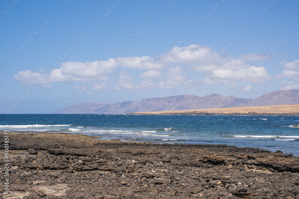 Seascape of the village of Caleta de Caballo, cliff and mountains of Famara in the background. Desertic landscape and ocean breaking against the rocks. Lanzarote, Canary Islands, Spain