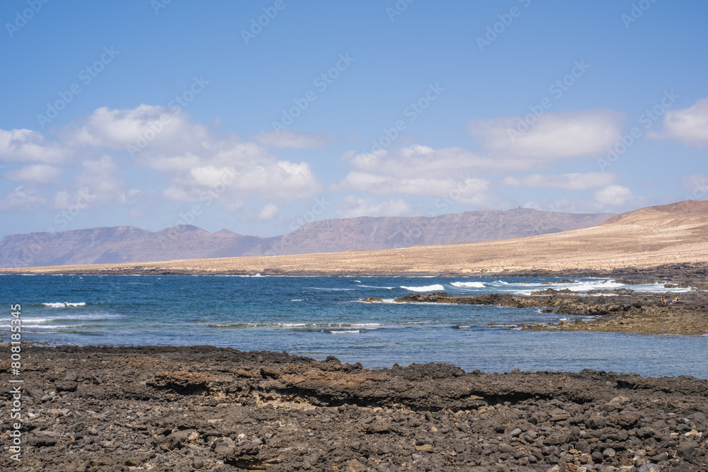 Seascape of the village of Caleta de Caballo, cliff and mountains of Famara in the background. Desertic landscape and ocean breaking against the rocks. Lanzarote, Canary Islands, Spain Seascape of the village of Caleta de Caballo, cliff and mountains of Famara in the background. Desertic landscape and ocean breaking against the rocks. Lanzarote, Canary Islands, Spain