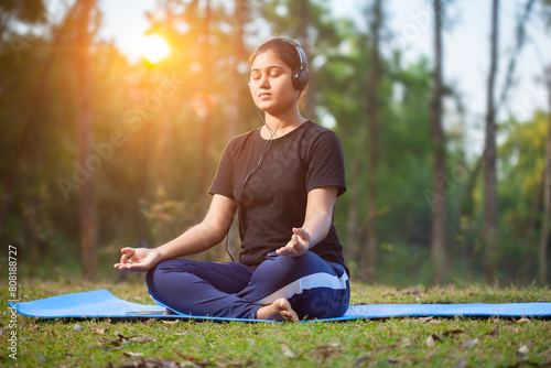 indian women practicing padmasana or lotus pose yoga with gyana mudra using headphone at park during morning sunrise