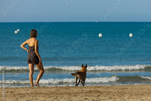 Back view of a woman and her dog standing at the water’s edge on a sandy dog friendly beach in the south of France on a hot summer’s day during a heat wave;.