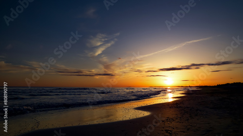 Beautiful sunset over a tranquil Mediterranean beach with golden sun reflected in shallow sea and on sparkling sand; amazing sunset with blue and orange sky and shallow waves on a sandy beach.