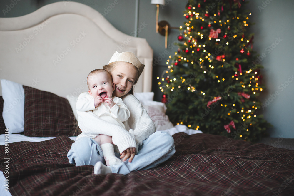 Smiling child girl 4-5 year old holding her baby sibling in bed over Christmas tree and glowing lights at home. Childhood. Winter holding season. Smiling child girl 4-5 year old holding her baby sibling in bed over Christmas tree and glowing lights at home. Childhood. Winter holding season.