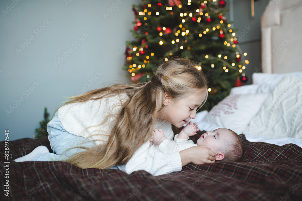 Smiling child girl 4-5 year old holding her baby sibling in bed over Christmas tree and glowing lights at home. Childhood. Winter holding season. Smiling child girl 4-5 year old holding her baby sibling in bed over Christmas tree and glowing lights at home. Childhood. Winter holding season.