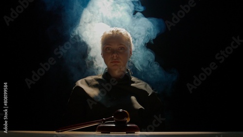 Female sitting at the desk with wooden hammer, dark room bright spotlight and smoke. Woman judge silhouette in the court room.