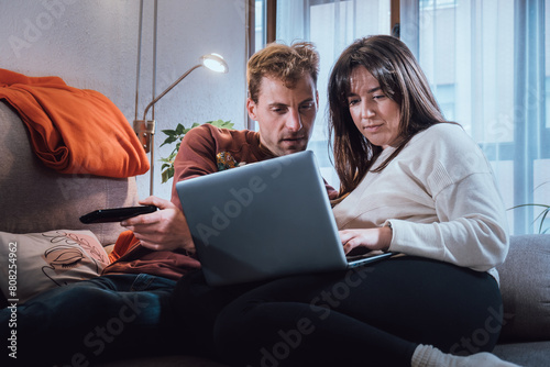 Young adult man and woman couple looking at a laptop on the couch. Real life people at home