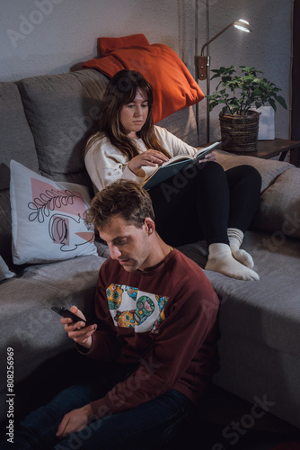 Young adult man and woman couple relaxing in the living room, the woman lying on the sofa reading a book and the man close to her looking at a smartphone. Real life people at home
