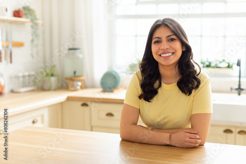A middle eastern woman is sitting at a wooden table in a white kitchen. The kitchen is bright with natural light coming in from a window.