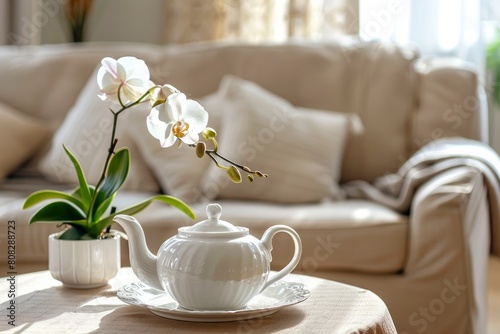 A white teapot and a pot with delicate orchid flower on coffee table with background of of a beige sofa in modern living room © Fatema