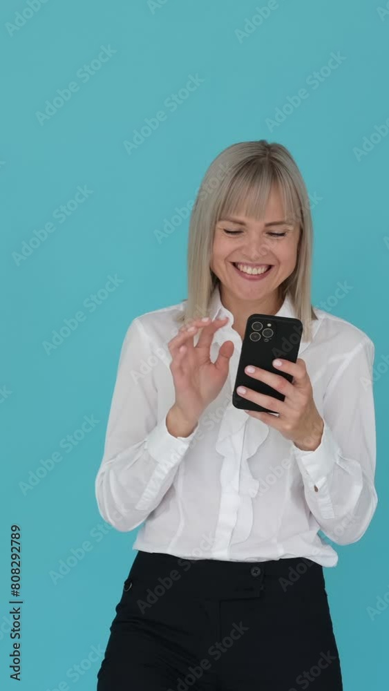 Joyful female office worker texting in online chat and laughing, spending leisure time with mobile phone on blue background. Vertical video. Joyful female office worker texting in online chat and laughing, spending leisure time with mobile phone on blue background. Vertical video.