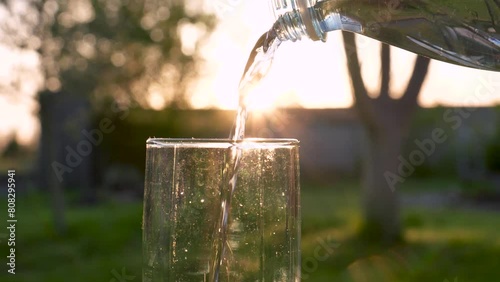 Glass of water in close up outdoors in green garden. Pouring pure mineral carbonated drinking water from bottle into glass against of sunset flooded sunlight. Healthy lifestyle and quenching thirst photo