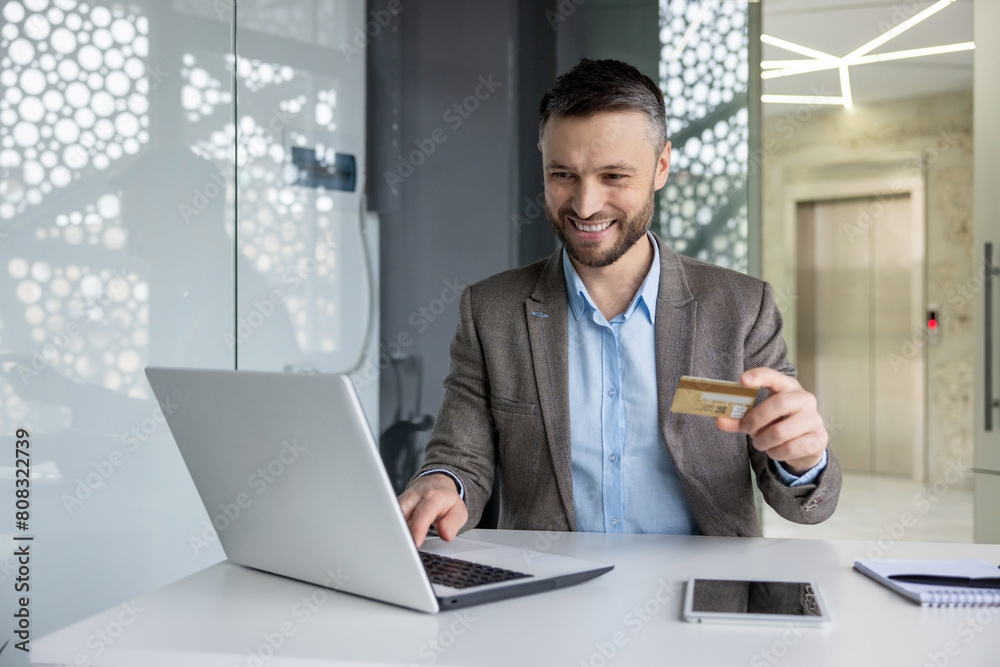 Cheerful businessman in a modern office space making an online purchase with a credit card and laptop. Professional and easy e-commerce transaction. Cheerful businessman in a modern office space making an online purchase with a credit card and laptop. Professional and easy e-commerce transaction.