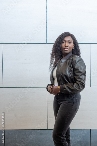 This image features a young Black woman standing confidently against a contemporary urban background of grey and white tiles. She wears a dark brown leather jacket over a light-colored top