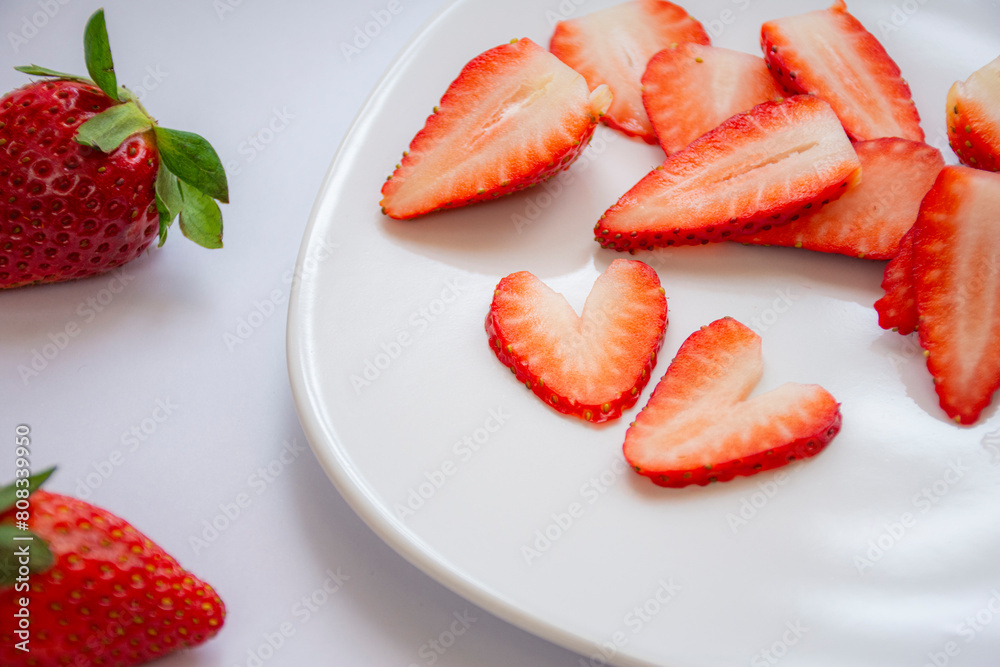 Two pieces of fresh red strawberry cut into a heart shape on a white plate with several more strawberries at the bottom Two pieces of fresh red strawberry cut into a heart shape on a white plate with several more strawberries at the bottom