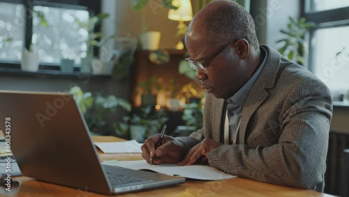Senior African American professor sitting at desk in library or office, reading information on laptop screen and taking notes in copybook photo