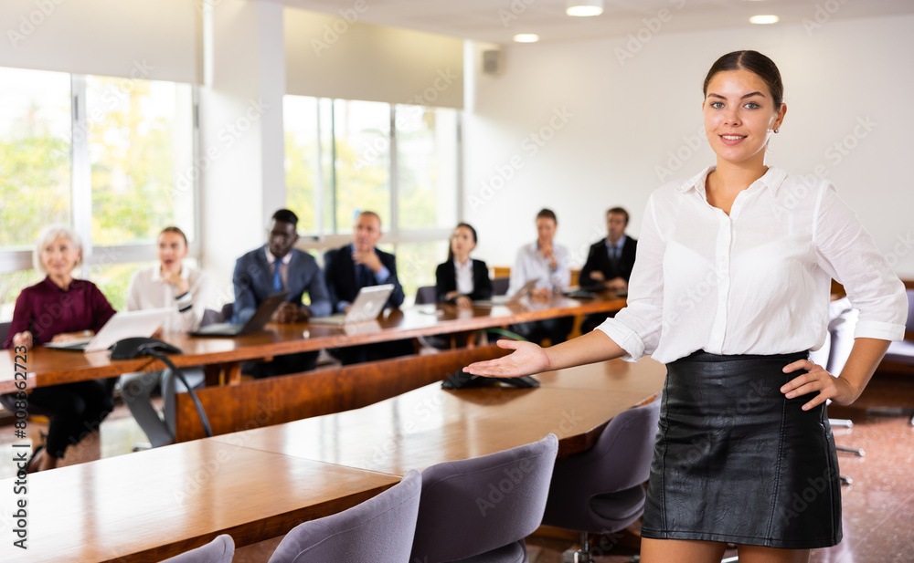 Friendly female office employee standing at workplace in meeting room, making inviting gesture
