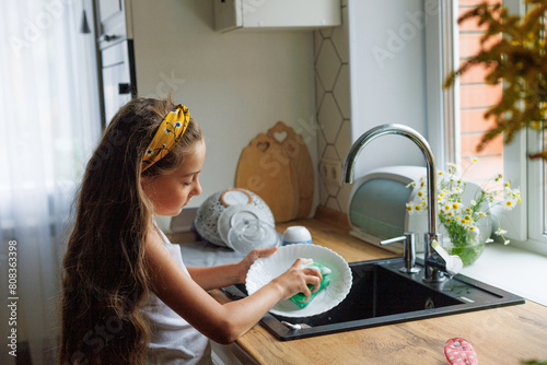 Murais de parede Cute little girl washing dishes with sponge in kitchen sink at home