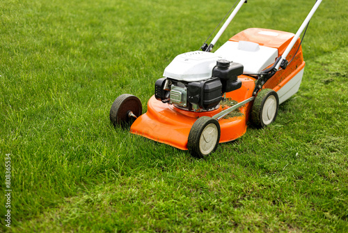 Top view of modern orange-grey gasoline lawn mower cutting bright lush green grass. Gardening work tools. Rotary lawn mower machine on lawn. Professional lawn care service. Place for text photo