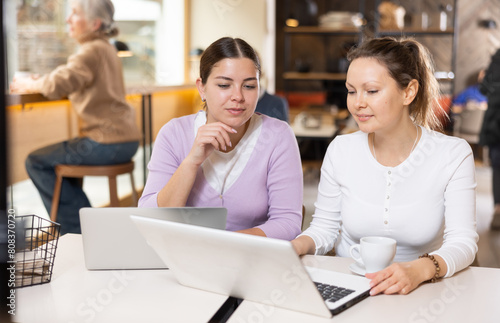 Two confident female cafe customers enjoying coffee while working on laptop