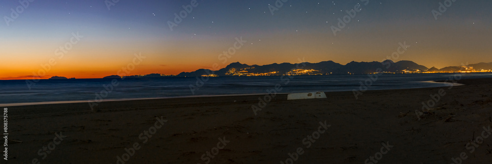 Panorama of sandy beach with white boat and illuminated Amalfi Coast during evening twilight after sunset with stars above, Paestum, Campania, Italy