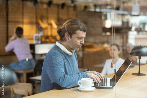 Concentrated male designer preparing new project on laptop, sitting in cozy cafe with cup of coffee