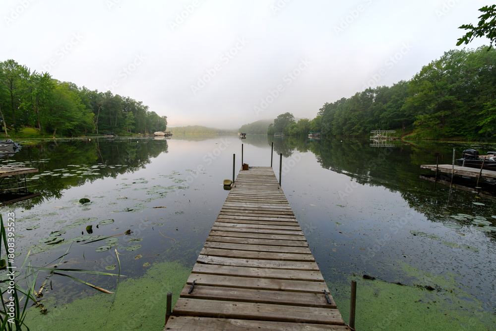 Naklejka premium Looking down a pier on a calm Wisconsin lake on a foggy morning. Fishing boat in distance.