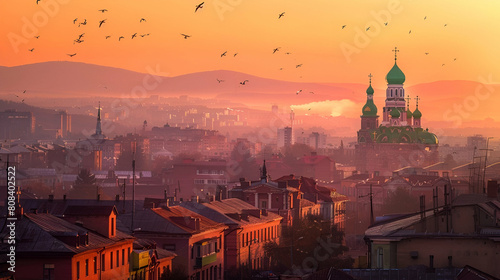 A panoramic view of the city skyline at sunset, with red roofs and an ornate domed church in the center. The sky is painted orange by setting sun, creating dramatic lighting over buildings