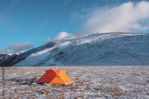 Orange tent on snow-covered stony pass in sunlight against snowy stone hill at early morning. Red tent in high mountains in freshly fallen snow. Snow-capped top and low clouds in blue sky at sunrise.