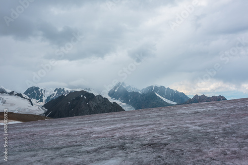 Dramatic vast misty top view from ice pass to big glacier tongue among sharp rocks and large snow-capped pinnacle in rainy low clouds. Dark atmospheric mountain silhouettes in rain in gray cloudy sky.