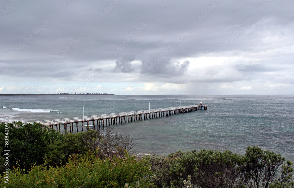 Wooden pier jetty extending into an ocean underneath a cloudy stormy sky at Point Lonsdale in Victoria, Australia