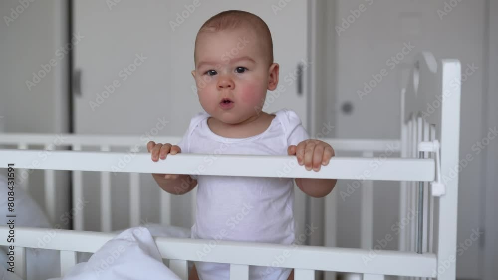 The baby is learning to stand. baby boy standing in the crib and holding on to the sides. Portrait of a newborn baby standing on his feet in a crib. Funny baby face. The baby is learning to stand. baby boy standing in the crib and holding on to the sides. Portrait of a newborn baby standing on his feet in a crib. Funny baby face.