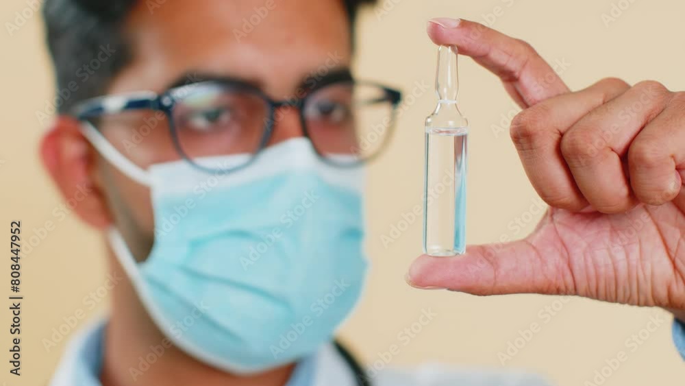 Indian doctor cardiologist man in protective medical mask holds an ampoule of vaccine medicine treatment injection. Laboratory testing. Arabian apothecary pharmacy guy isolated on beige background Indian doctor cardiologist man in protective medical mask holds an ampoule of vaccine medicine treatment injection. Laboratory testing. Arabian apothecary pharmacy guy isolated on beige background