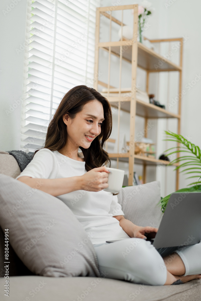Woman takes pleasure in sipping her coffee as she focuses on her laptop, comfortably seated on a couch in a bright home setting. Woman takes pleasure in sipping her coffee as she focuses on her laptop, comfortably seated on a couch in a bright home setting.