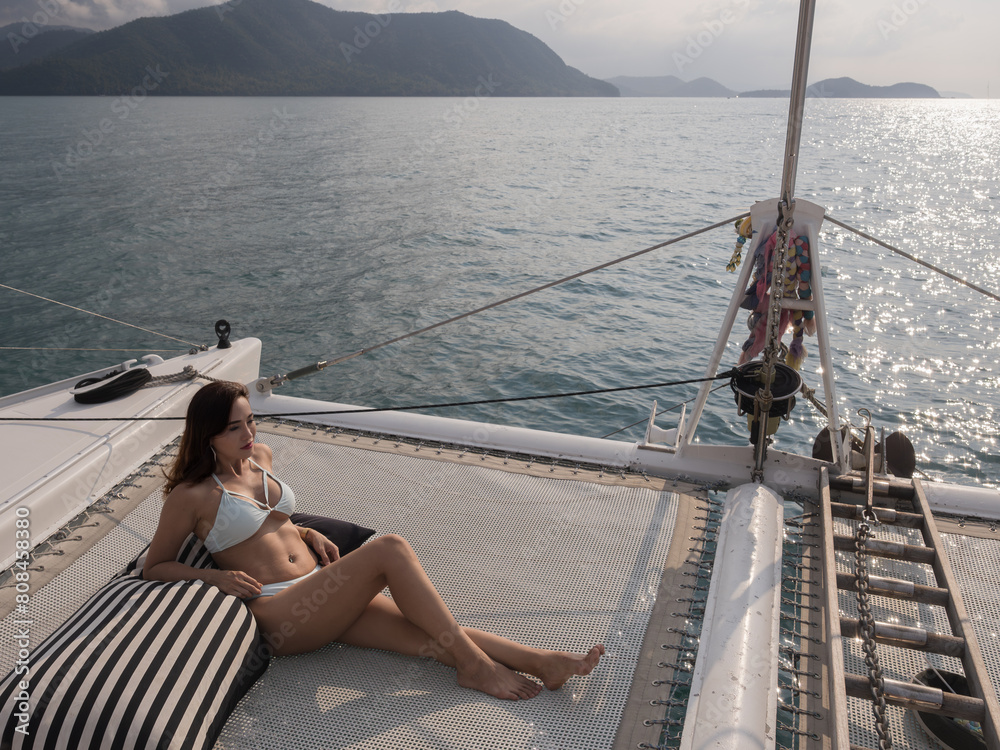 Happy Asian woman in white bikini relaxing, sitting on the polyester net of a luxury yacht during a sailing trip on the sea, enjoying the summer holiday concept
