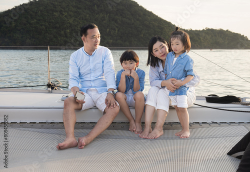 Happy Asian family relaxing, sitting, and playing on the polyester net of a luxury yacht during a sailing trip on the sea, enjoying the summer holiday concept.