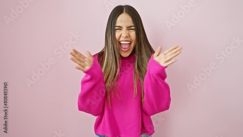Excited young latin woman in sweater celebrates lunatic success. standing, arms raised, mad with joy over a pink isolated background. crazy victory expression of a beautiful winner! photo