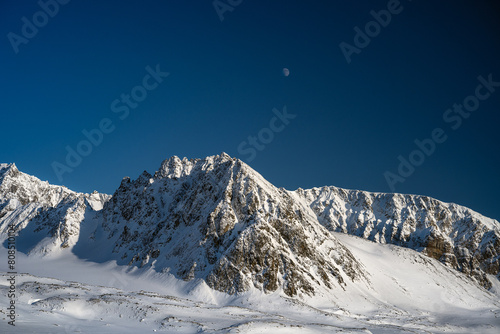 2023-13-31 SNOW COVERED JAGGED PEAKS WITH A BLUE SKY AND A MOON NEAR SVALBARD NORWAY IN THE ARCTIC