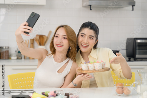 Two Asian women in a modern kitchen taking a selfie with a plate of beautifully decorated cakes, both smiling happily.
