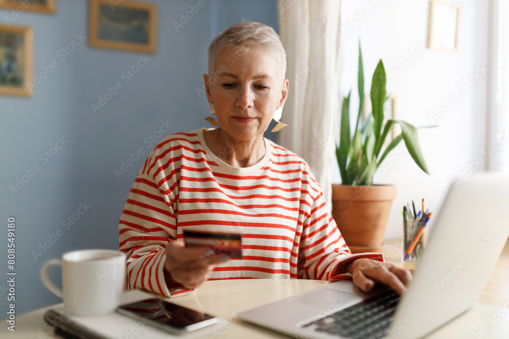 Selective focus of female hand holding credit card doing online payment with happy smiling face sitting at table with smartphone and mug, using laptop, buying plane ticket or travel tour for weekend Selective focus of female hand holding credit card doing online payment with happy smiling face sitting at table with smartphone and mug, using laptop, buying plane ticket or travel tour for weekend