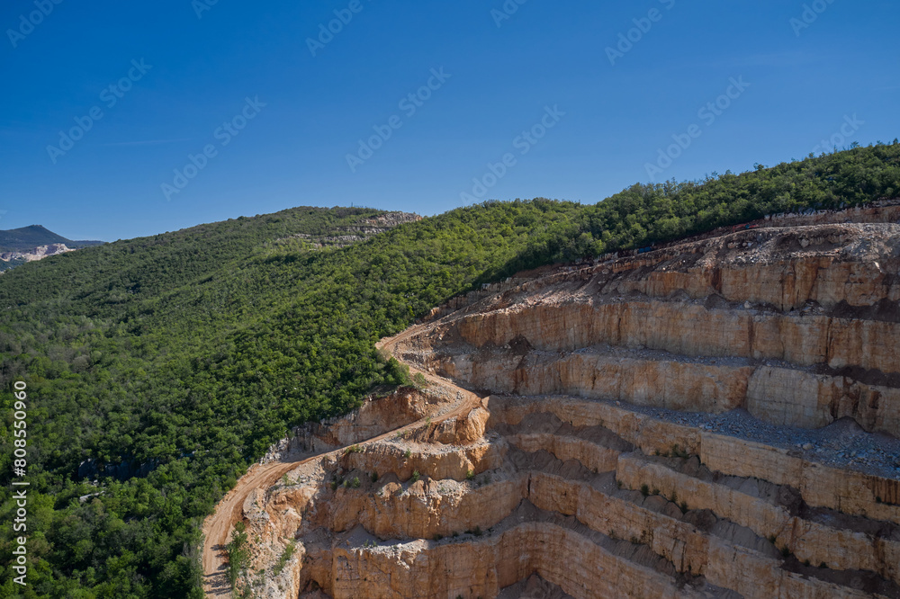 Steps of a marble quarry in the background green forest blue sky. Open pit marble extraction. Large marble quarry against the blue sky.